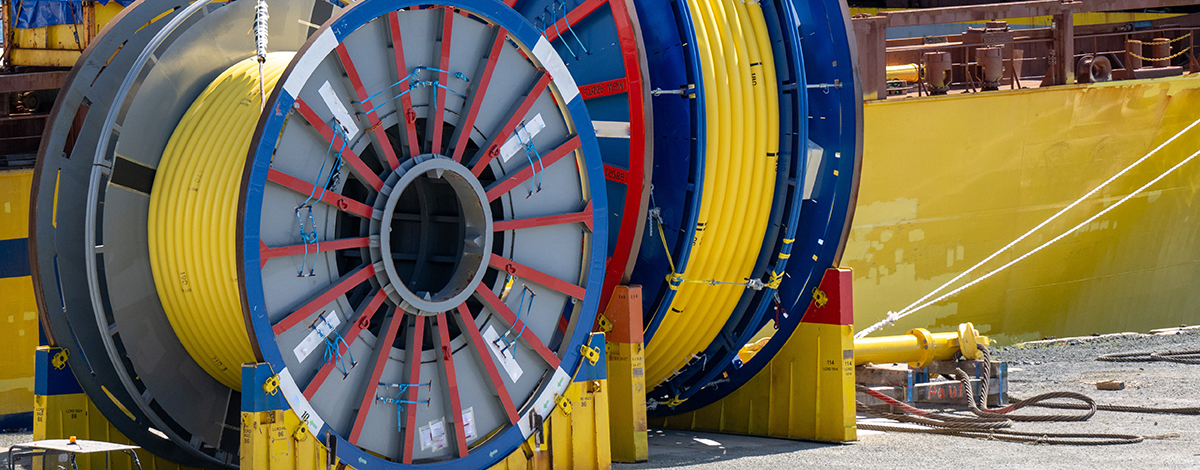 Bulk sub-sea industrial glass fiber optic cable on a metal spool on a ship's stand. The yellow data line is coiled around a black reel in a storage yard. 
